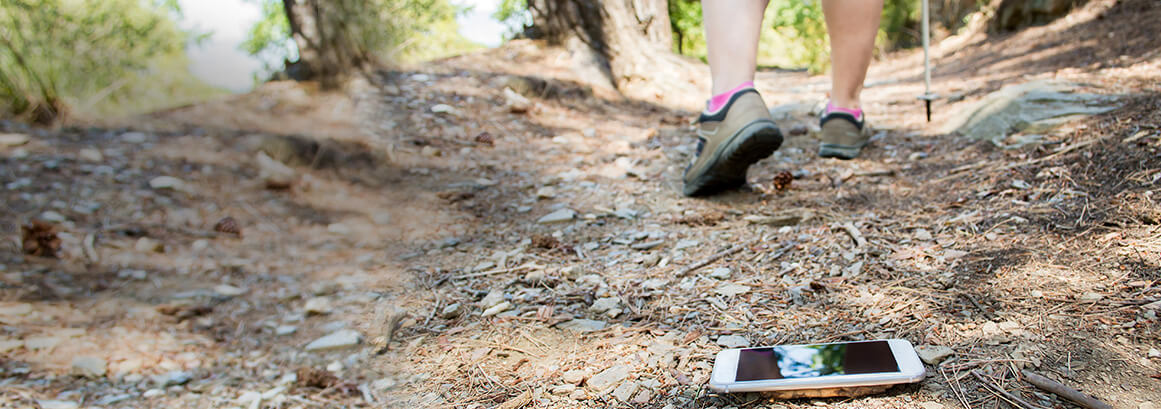 Auf dem Waldboden liegt ein das Handy das einer Person aus der tasche gefallen ist. Von der Person sieht man nur noch die Füße und Wanderstöcke in der Ferne.